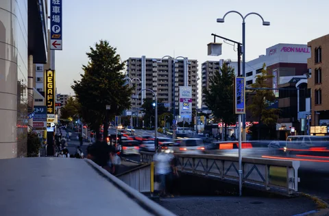 Red vehicle light trails cross an urban road beside a railing and twin curved streetlights.