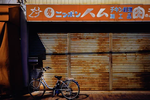 A bicycle rests before rust-streaked shutters beneath a long orange shop sign and diagonal shadows.