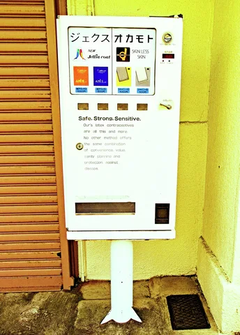 A small white vending machine stands on a pedestal beside a brown shutter and yellow wall.