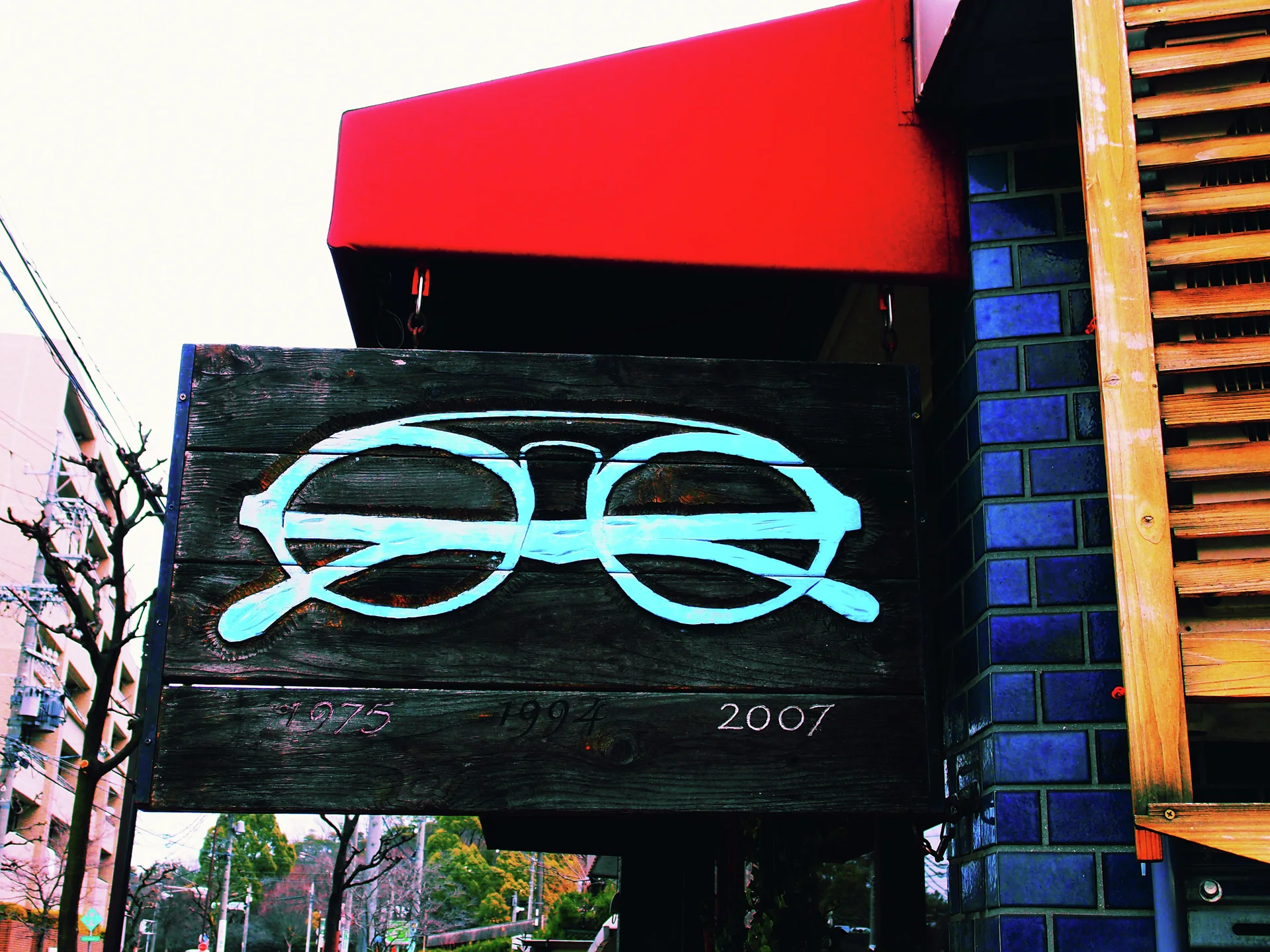A turquoise eyeglasses graphic is painted on a dark wooden hanging sign beneath a red awning.