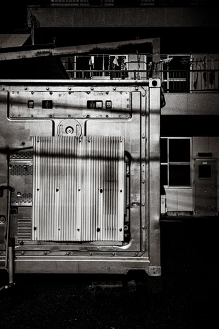 A large rectangular metal door with heavy hinges fills a shadowy industrial interior in monochrome.