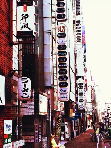 Tall stacked business signs crowd a building-lined sidewalk beside a hanging lantern and distant pedestrians.