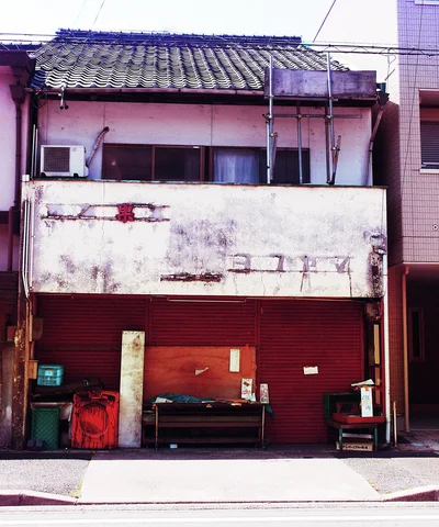 A weathered two-story facade has a tiled roof above red boarded panels and assorted items on the pavement.