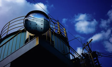 A round convex mirror reflects a pale building above yellow rooftop railings beneath blue sky and clouds.