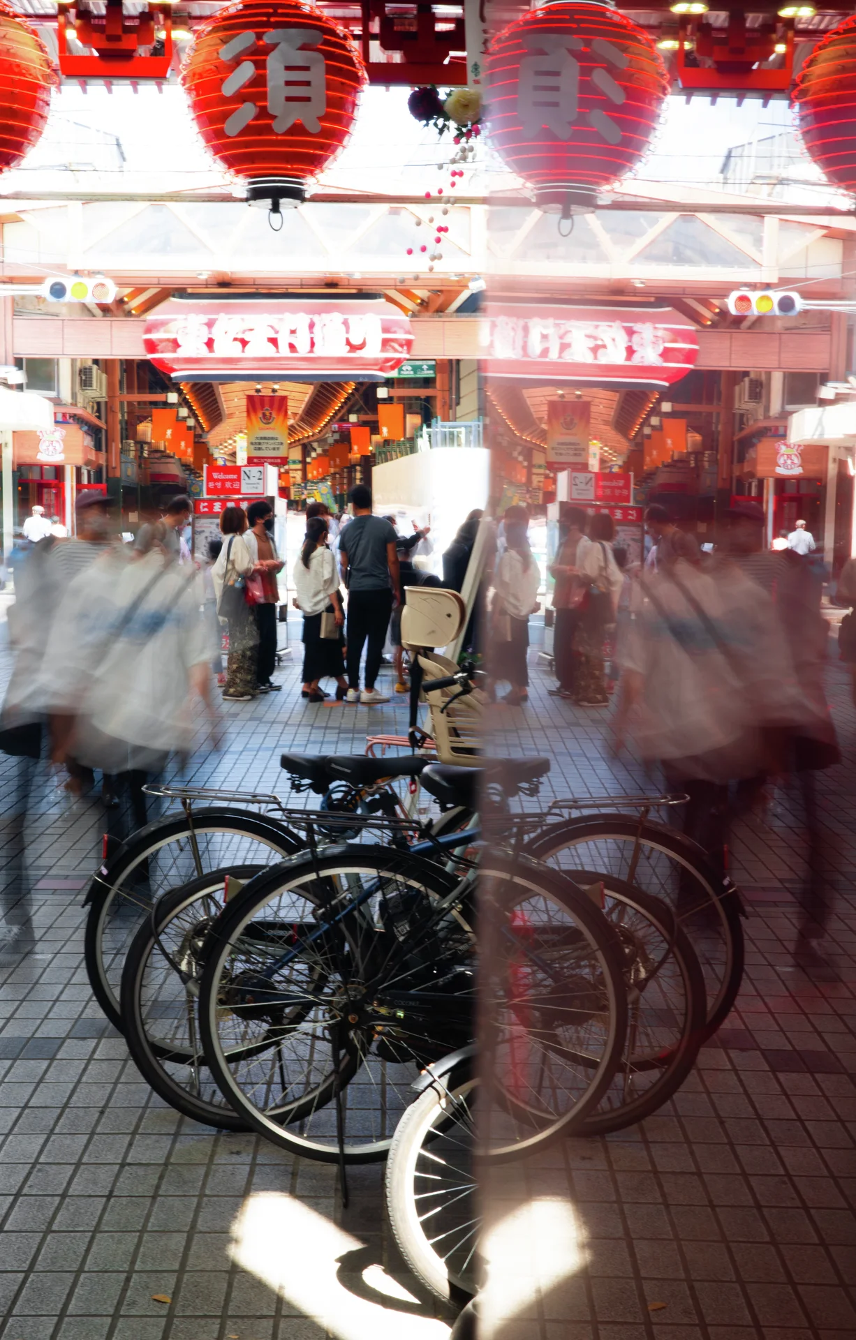 Bicycles and blurred reflections fill the foreground beneath red lanterns at a busy covered passage.