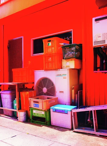 Crates and containers surround an outdoor air conditioner against vivid red siding beneath narrow windows.