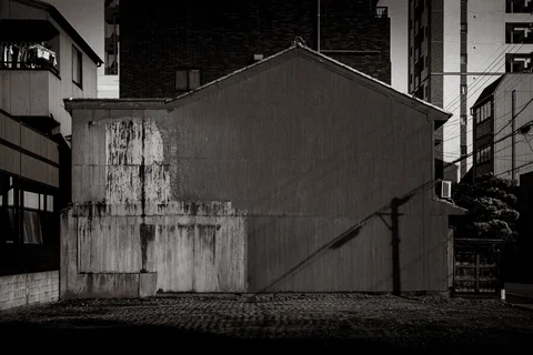 A stained blank gabled wall stands between taller buildings above patterned paving in black and white.