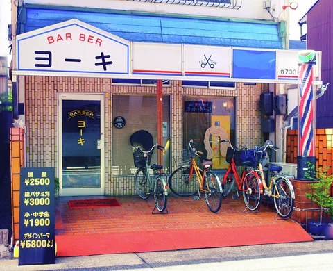 Several bicycles stand outside a blue-trimmed barber shop beside a striped pole and freestanding price board.
