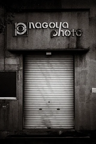 Raised Nagoya photo lettering sits above a closed shutter on a weathered monochrome facade.