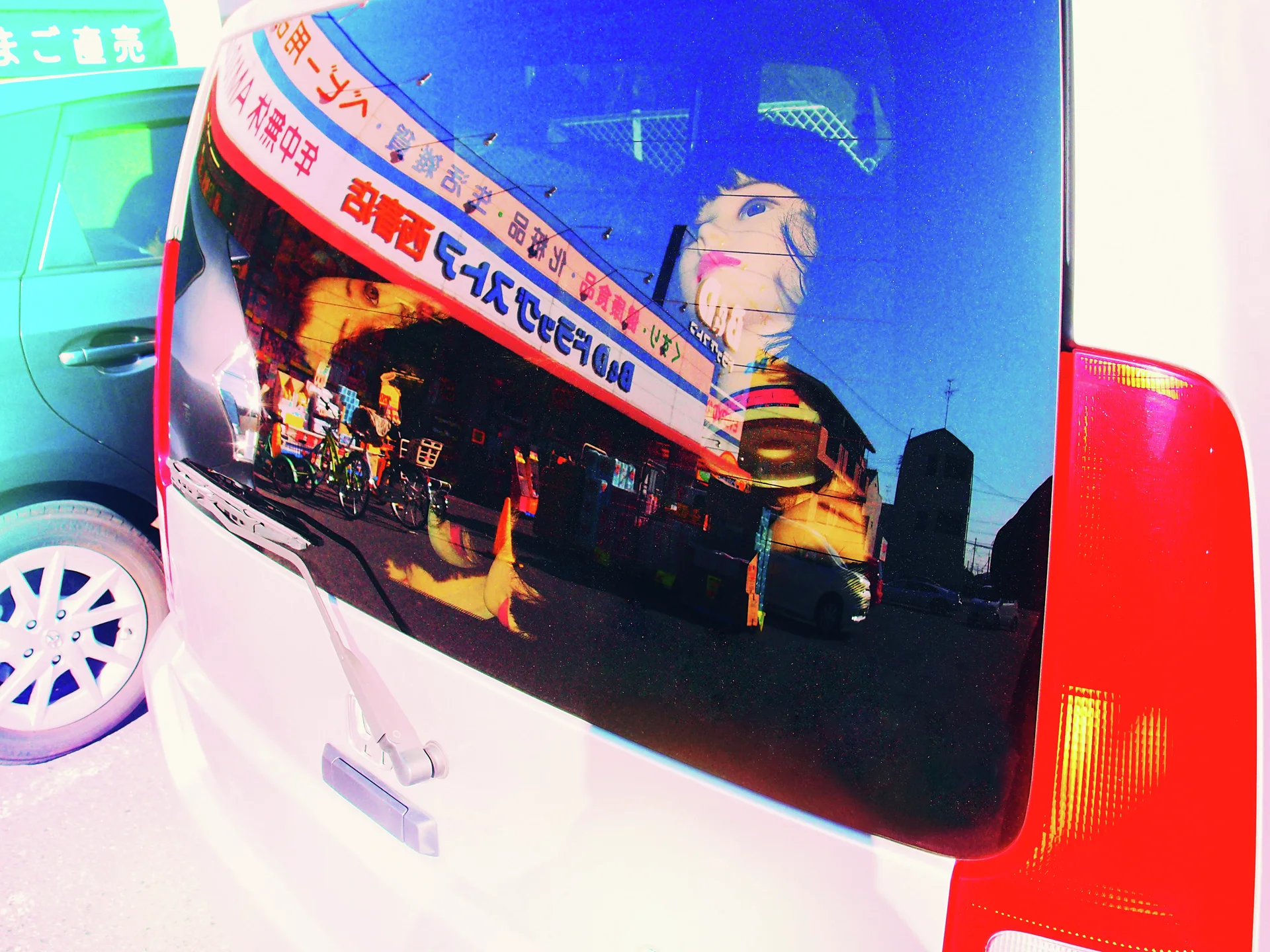 A white vehicle's rear window reflects colorful shop signs and a large illustrated face beside its red taillight.