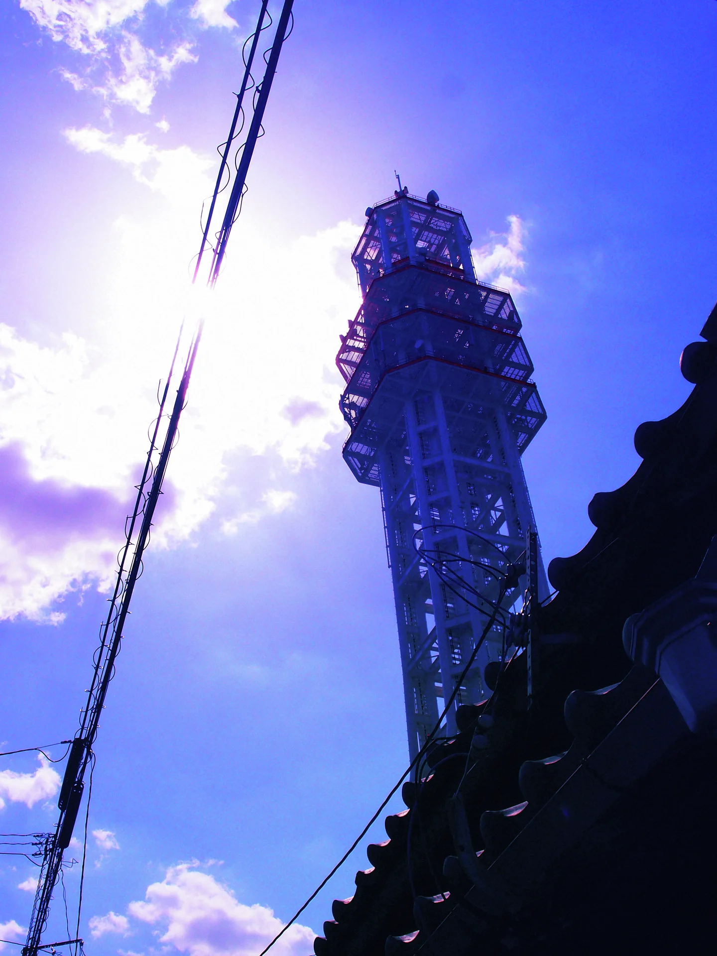A metal lattice tower rises beside a dark roof edge and utility wires beneath bright blue clouds.
