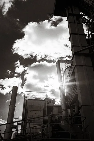Sunlight breaks around an industrial structure beneath bright clouds in a high-contrast black and white view.