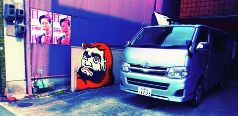 A silver van parks beside a red round-faced figure display and two portrait posters.
