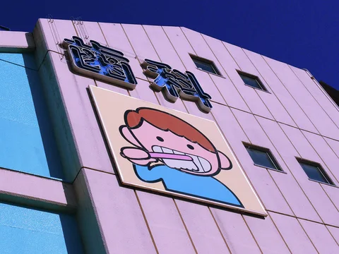 A cartoon child brushing teeth decorates a pale pink building beneath raised Japanese dental lettering.