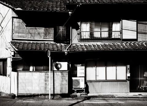 A weathered two-story house has tiled eaves, closed windows, and stacked air conditioners in black and white.
