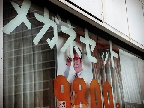 Large pale Japanese lettering and orange price numerals overlay a printed portrait behind a curtained shop window.