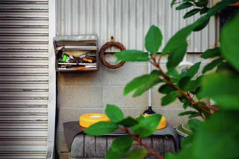 Green leaves partly obscure a yellow basin below a wall-mounted shelf and coiled hose.
