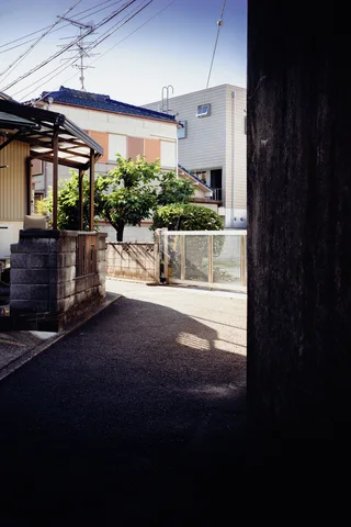 A dark passage frames a sunlit residential lane with low walls, shrubs, and overhead wires.