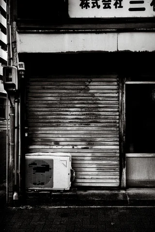 A rust-marked closed shutter stands behind an outdoor air conditioner beneath a shop sign in black and white.