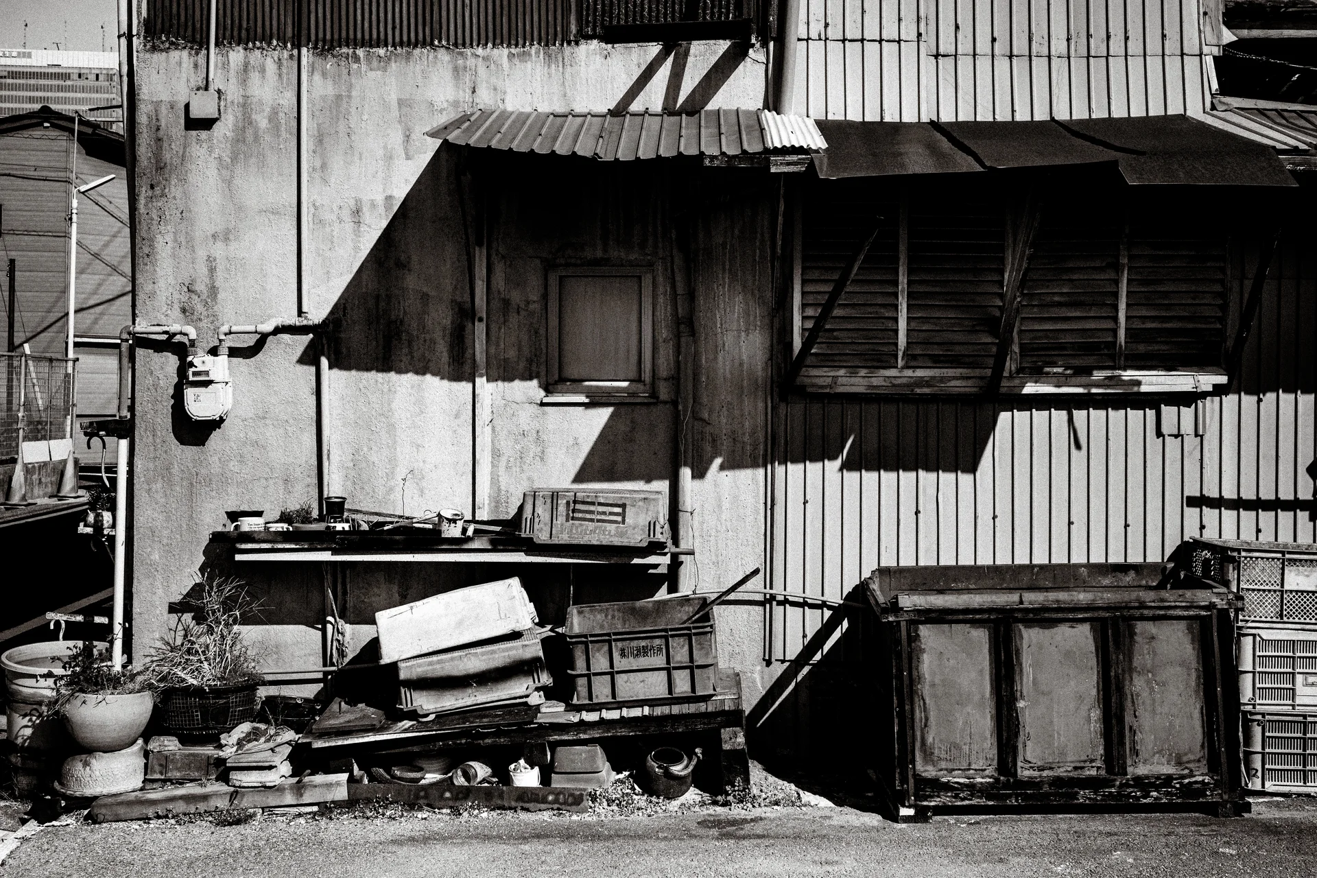 Discarded boxes and containers rest against a weathered corrugated building beneath projecting window awnings in black and white.