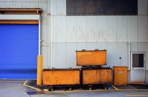 Rusty orange containers stand against a corrugated industrial wall beside a vivid blue shutter and narrow door.