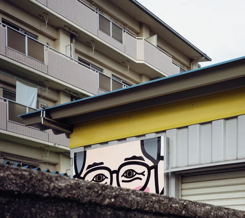 A painted face peers above a rough wall beneath a yellow fascia and apartment balconies.