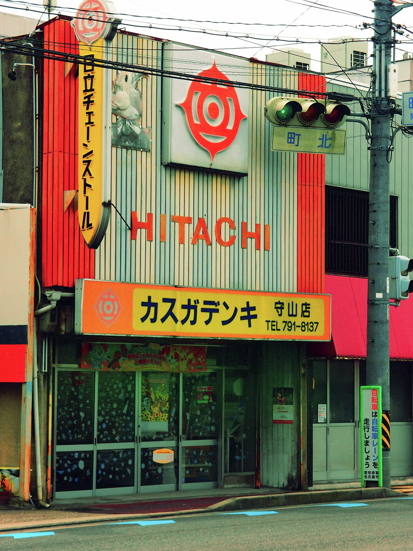 Red Hitachi lettering covers a corrugated storefront above a yellow sign beneath utility wires and traffic lights.