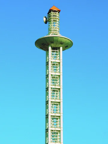 A tall lattice tower with a broad circular platform and orange cap rises against clear blue sky.