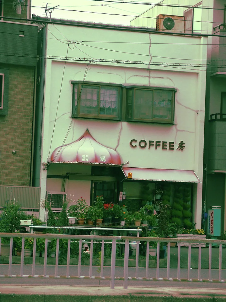 A small pale coffee shop has dark upper windows and a pink domed entrance canopy above greenery.