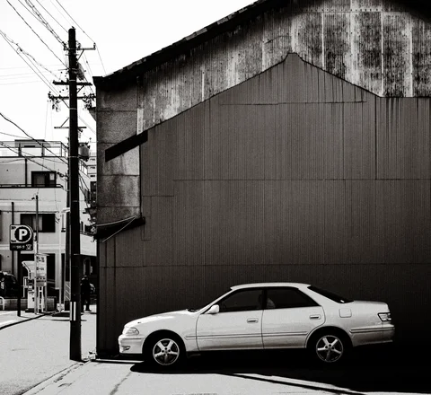A white sedan sits beside a large blank corrugated building wall in monochrome.