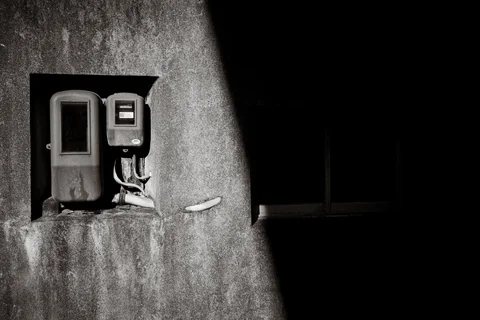 Utility boxes and exposed cables catch sunlight beside a shadowed window on a rough wall in black and white.