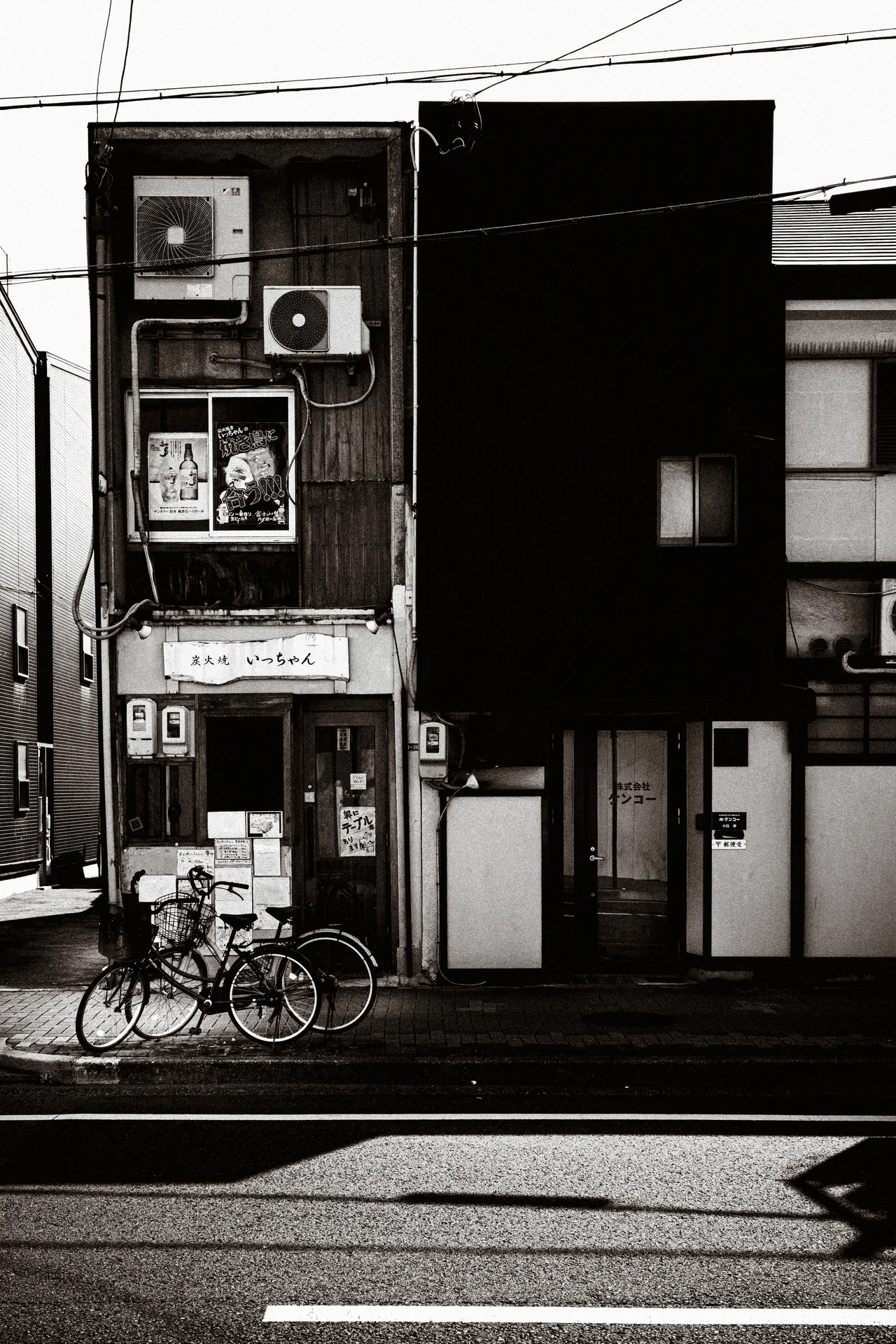 Bicycles stand outside a narrow building with air conditioners and contrasting dark facade panels in monochrome.