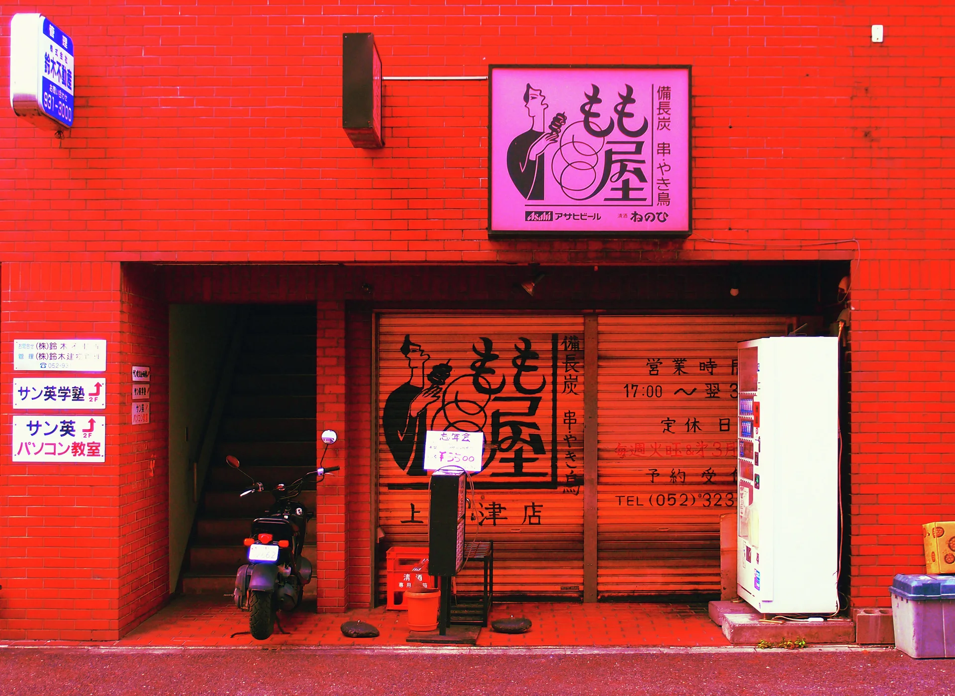 A red storefront surrounds orange shutters with Japanese lettering beside a scooter and white vending machine.