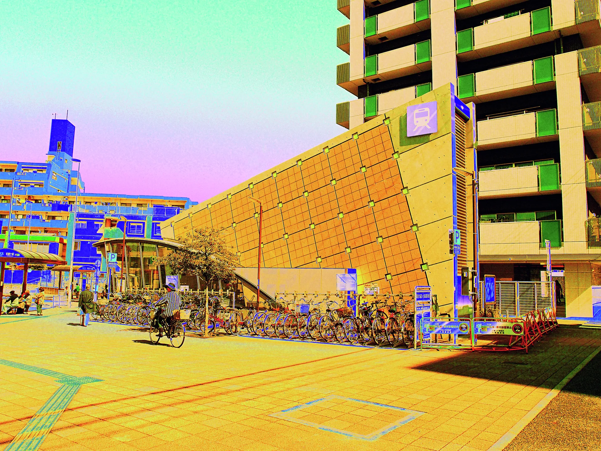 Rows of parked bicycles line a yellow station wall beside a tall apartment building and broad tiled plaza.
