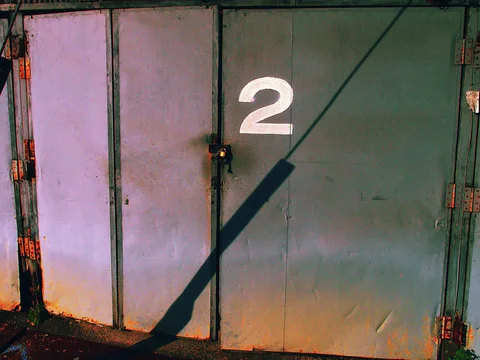 A padlocked metal door marked with a white 2 is crossed by a long diagonal shadow.
