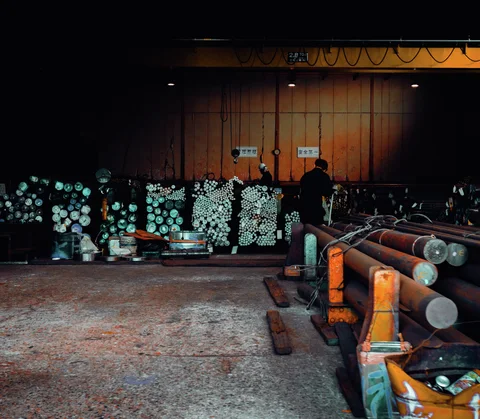 Bundles of cylindrical metal stock fill an open workshop, with thick bars supported on orange stands.