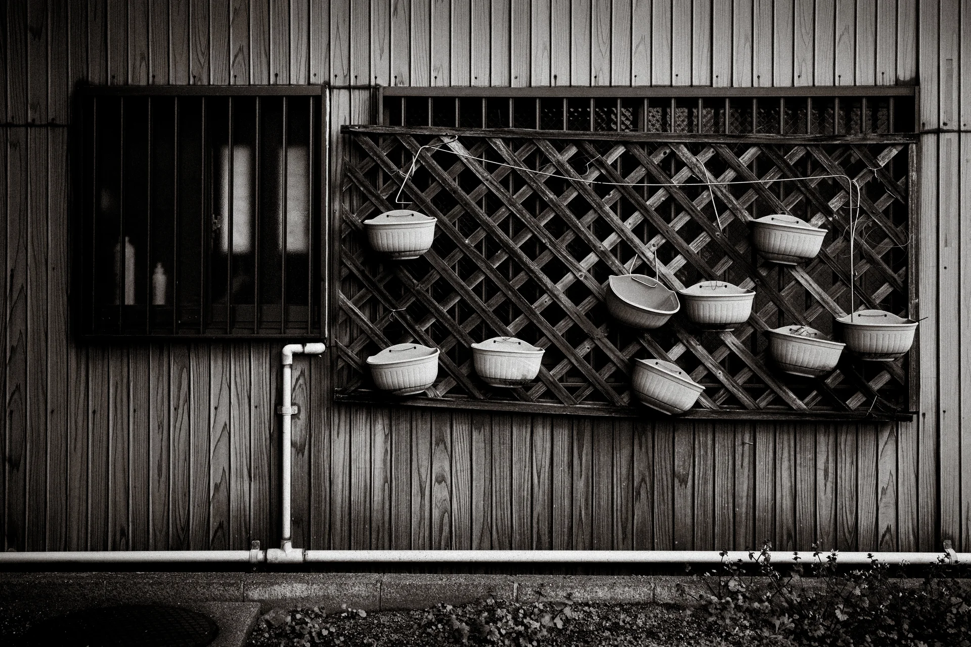 Empty white hanging pots dot a wooden trellis beneath barred windows in black and white.