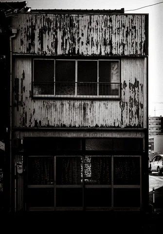 A peeling two-story corrugated facade has dark upper windows and a glazed ground-floor entrance in black and white.