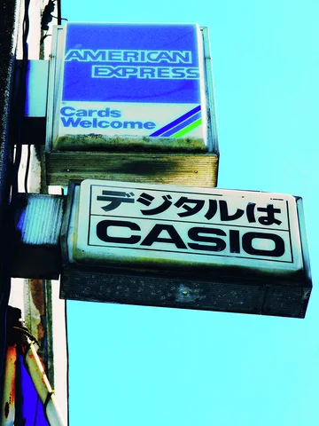 American Express and Casio signs project one above the other against a bright turquoise sky.