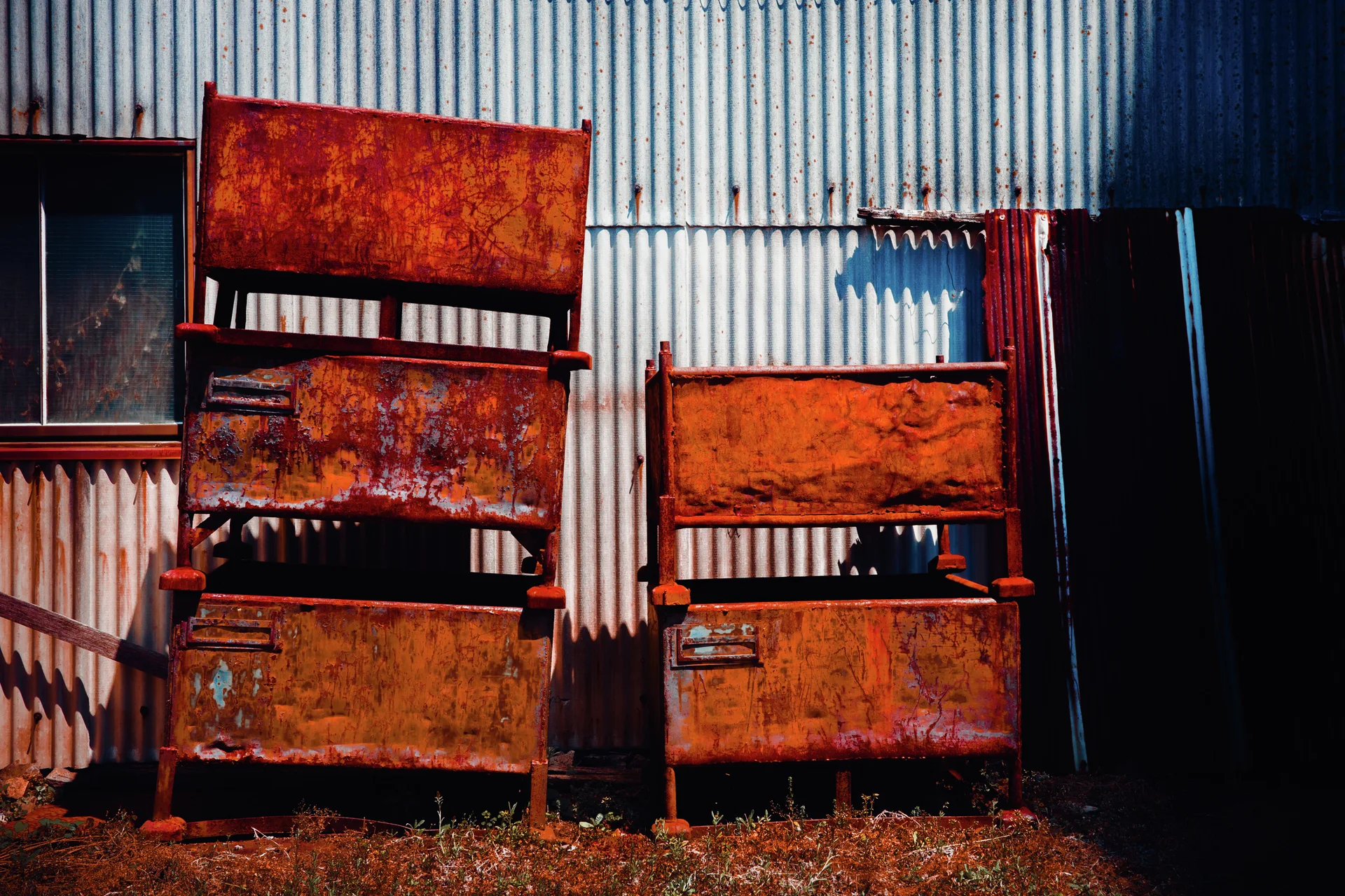 Stacks of heavily rusted rectangular metal containers stand before a pale corrugated wall.