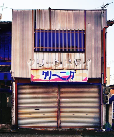 A weathered cleaning-shop sign hangs above closed shutters beneath a barred upper window.