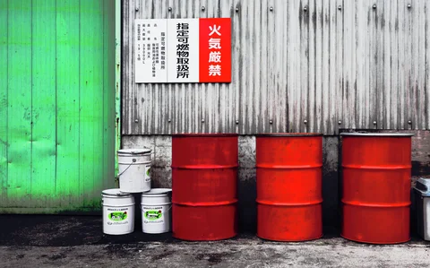Three red drums and stacked white buckets stand against corrugated siding beside a large green door.