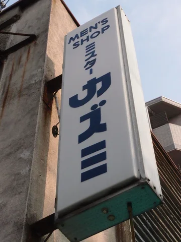 Blue Japanese characters run down a projecting white MEN'S SHOP sign on a weathered wall.