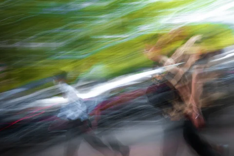 A heavily blurred pedestrian and vehicles form diagonal streaks beneath green roadside trees.