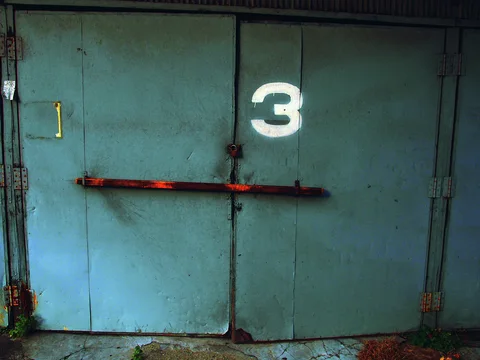 A white 3 marks blue metal double doors secured by a long horizontal rusted bar.