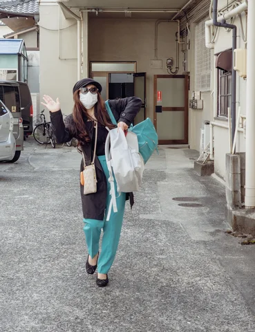 A masked person in turquoise trousers carries bags along a narrow lane and raises one hand.