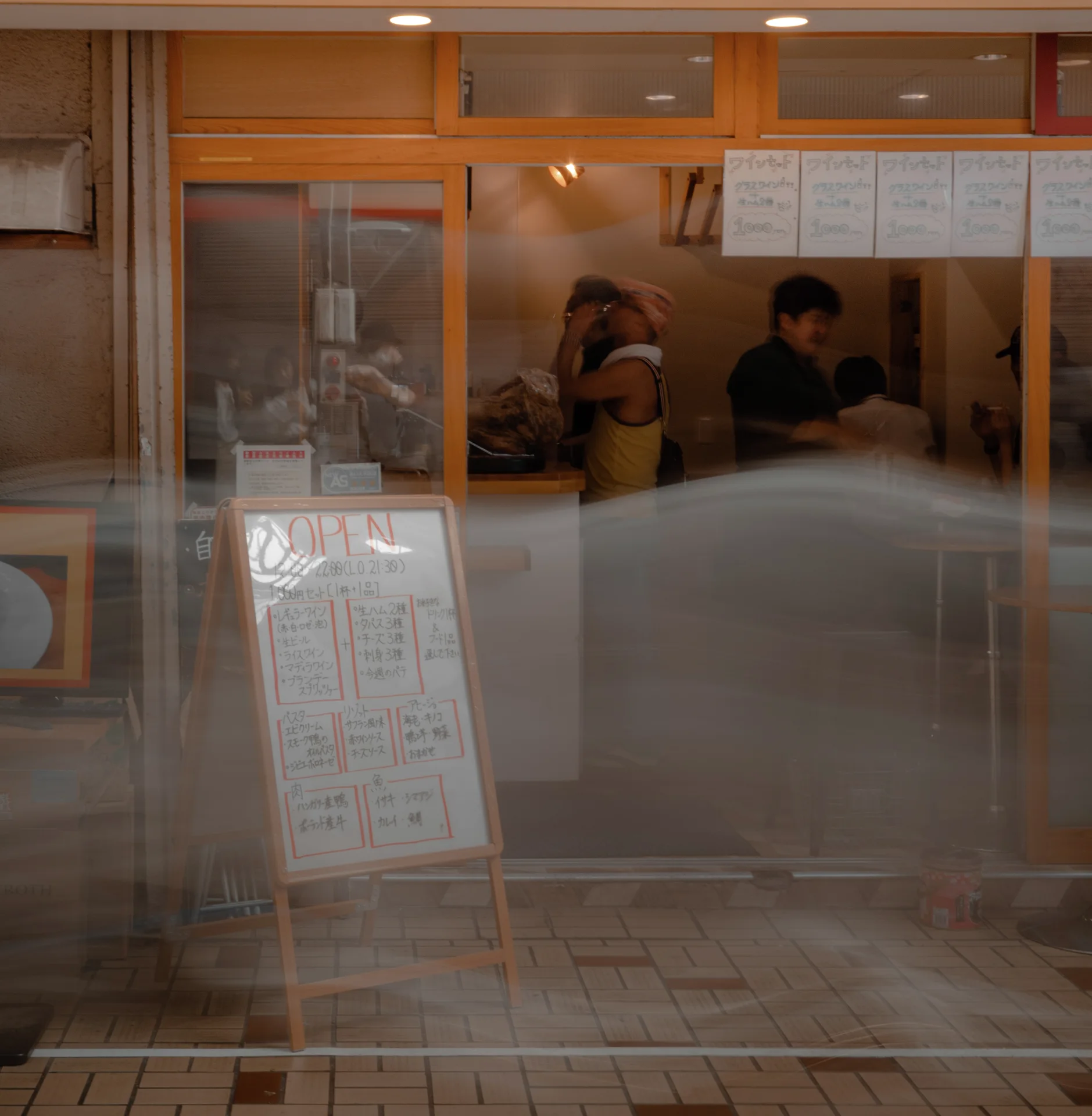 Street reflections overlay people inside a glass-fronted shop with orange window frames and a standing menu board.