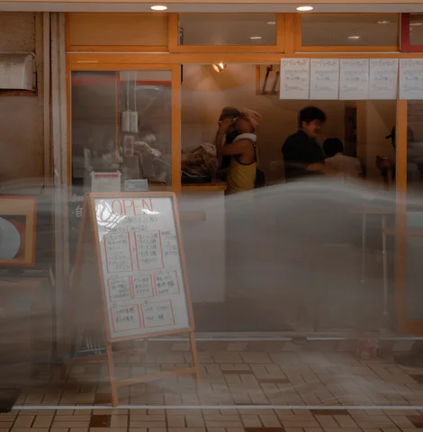 Street reflections overlay people inside a glass-fronted shop with orange window frames and a standing menu board.