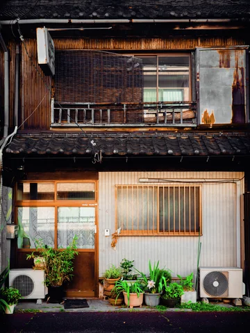Potted greenery clusters between two air conditioners beneath wooden doors and a weathered upstairs balcony.
