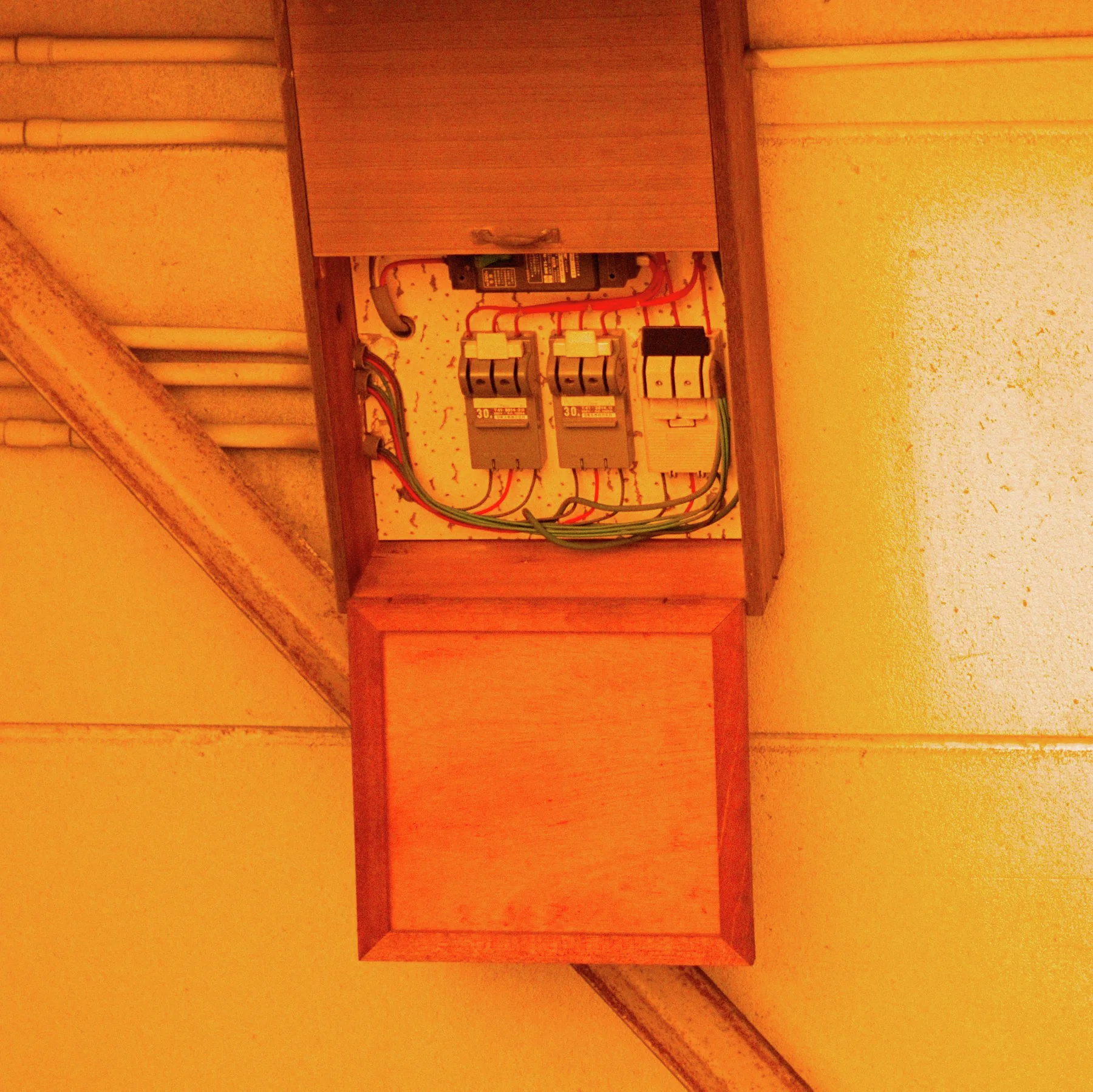 An open electrical box exposes switches and tangled wires against an orange-lit wall beside conduit.
